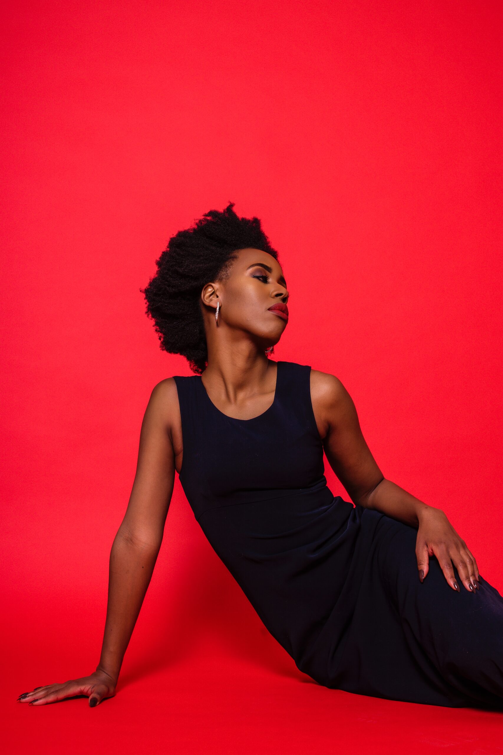 Side profile of Elle Friberg wearing her afro, a navy blue dress and red lipstick against a red backdrop