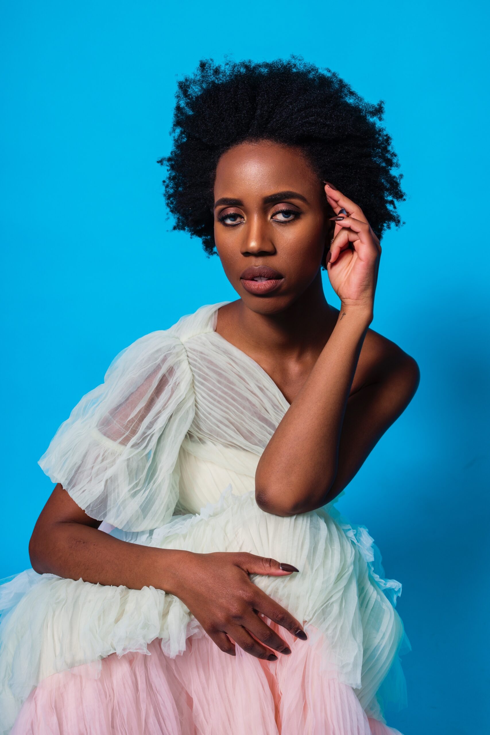 Elle looking directly at the camera in a pastel floor length dress against a blue  backdrop, sitting down